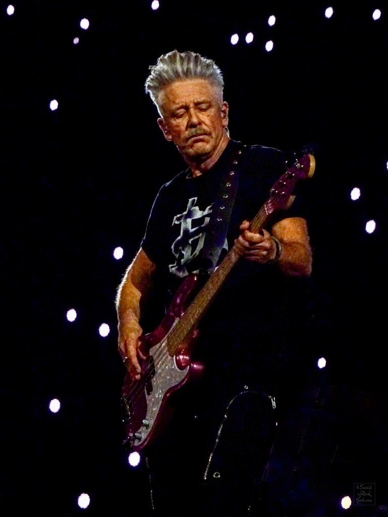A close up photo of Adam Clayton from U2 playing bass guitar.  White dots projected on the screen surround him. From the U2:UV Achtung Baby at the Sphere concert in Las Vegas March 1, 2024. 