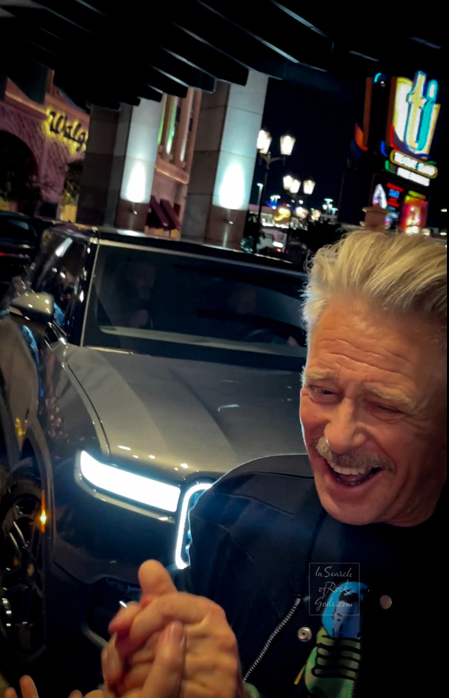 A photo of Adam Clayton smiling and shaking hands with a fan in a crowd of people outside of the Zoo Station exit at the Venetian Resort and Casino.  Taken on February 29, 2024
